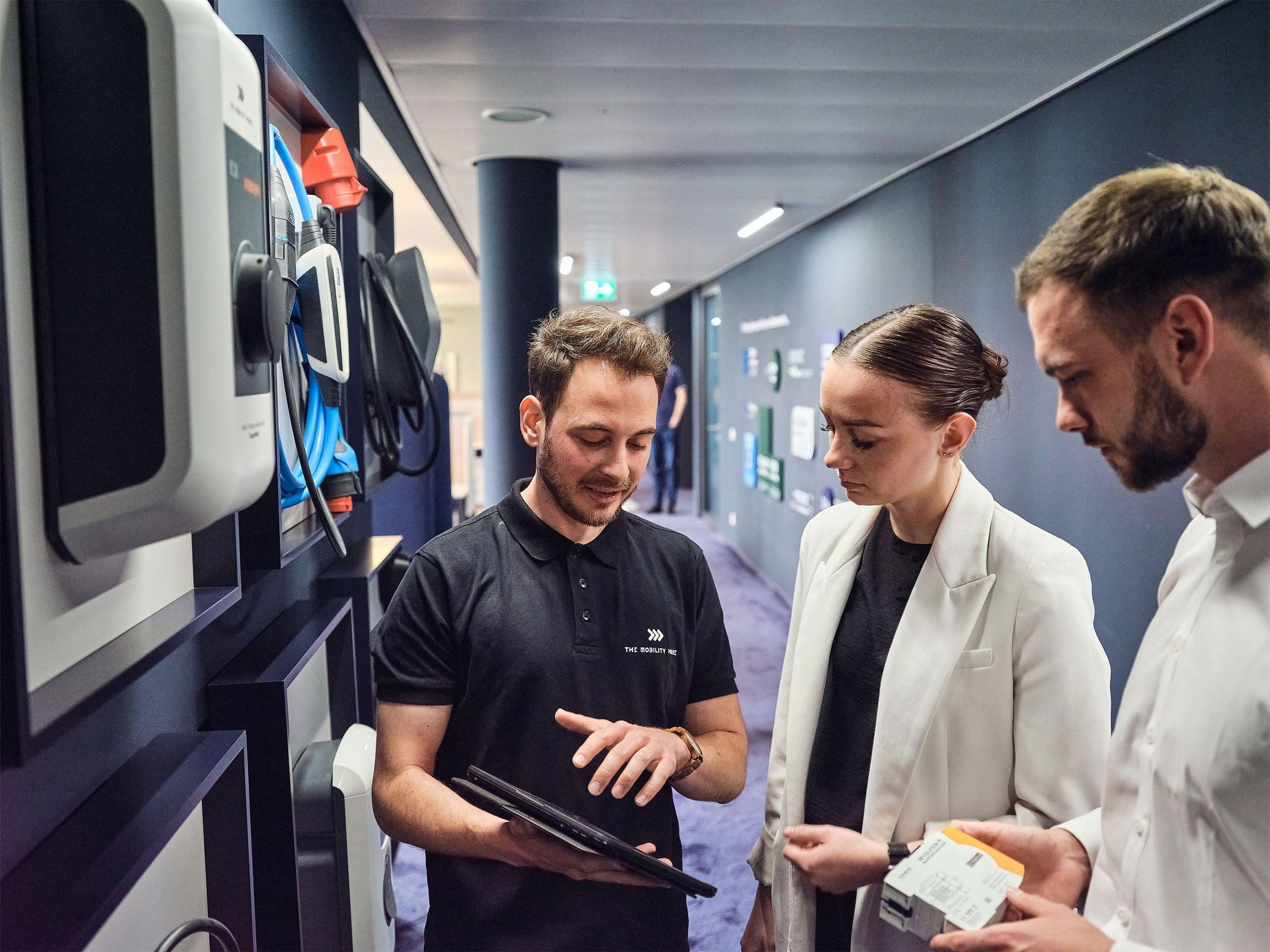 3 Personen fuehren ein Gespraech vor einer Wand mit Ladestationen